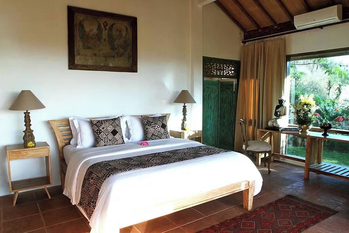 A bedroom with a wooden bed, two bedside lamps, a green door, a window with a view of greenery, and a table with a vase of flowers.