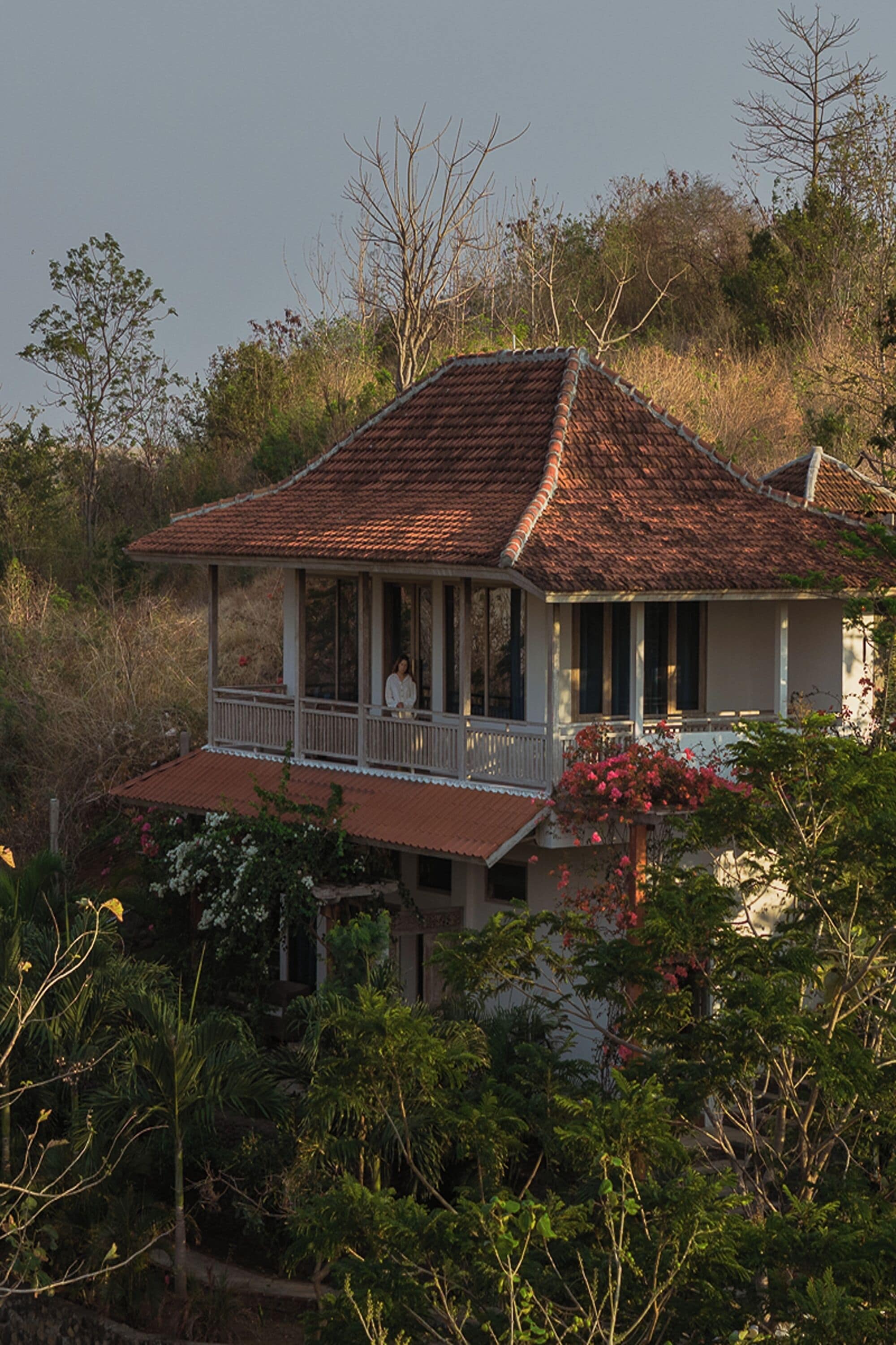 Une maison à deux étages dotée d’un balcon, entourée d’une végétation luxuriante et d’arbres.