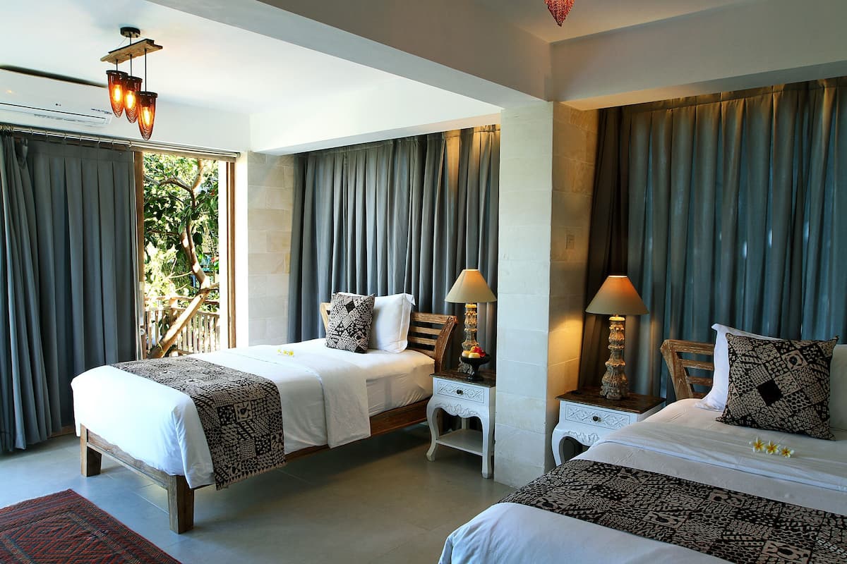 A hotel room with two beds, a nightstand with a lamp, a window with curtains, and a view of greenery outside.