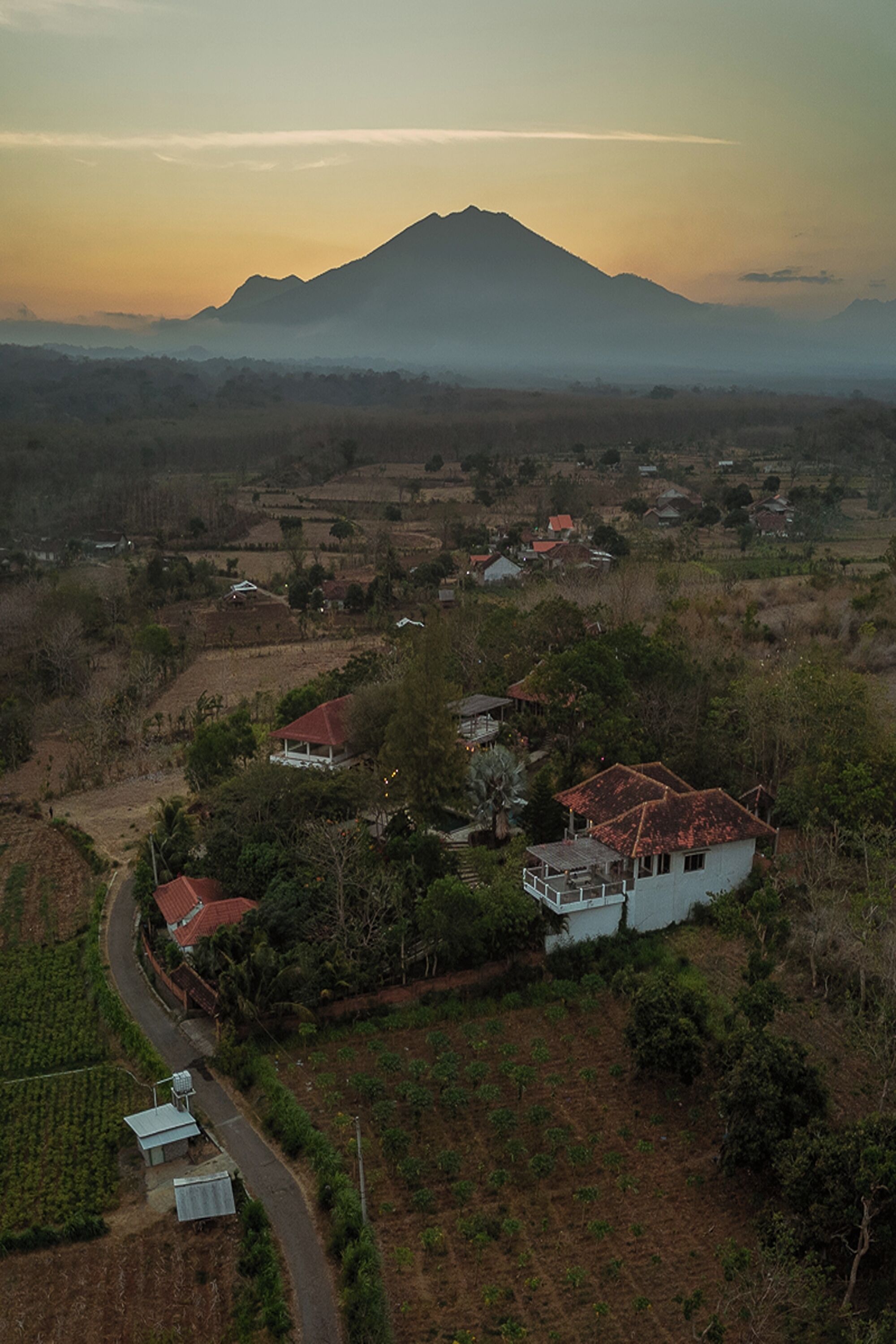 An aerial view of a rural area with a prominent mountain in the background, surrounded by fields and scattered houses with red roofs.