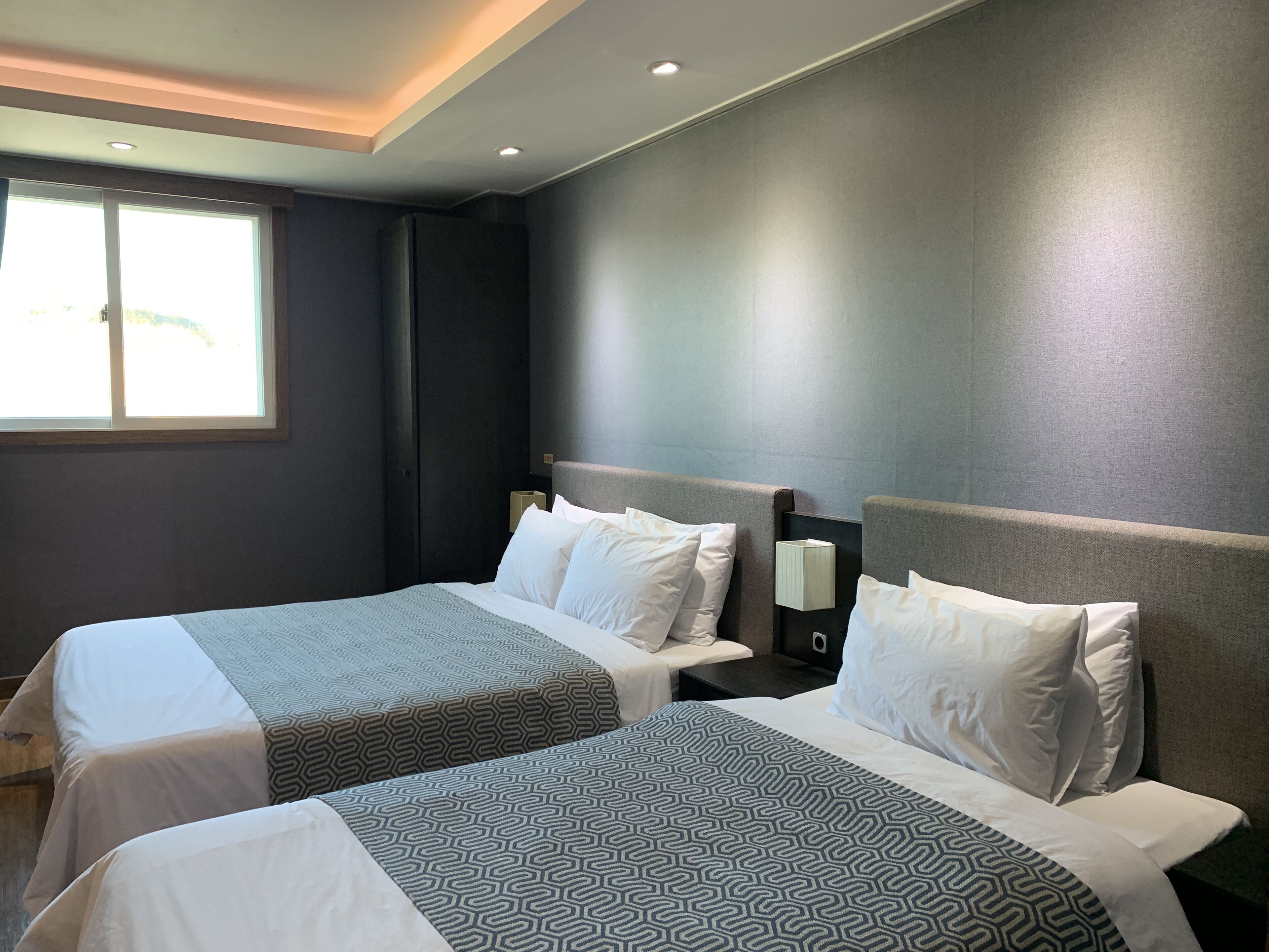 Habitación de hotel con dos camas, cada una con una colcha estampada, una mesita de noche con lámpara y una ventana que proporciona luz natural.