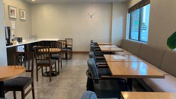Un comedor moderno con mesas y sillas de madera, una encimera de cocina con electrodomésticos, y un ventanal que proporciona luz natural.