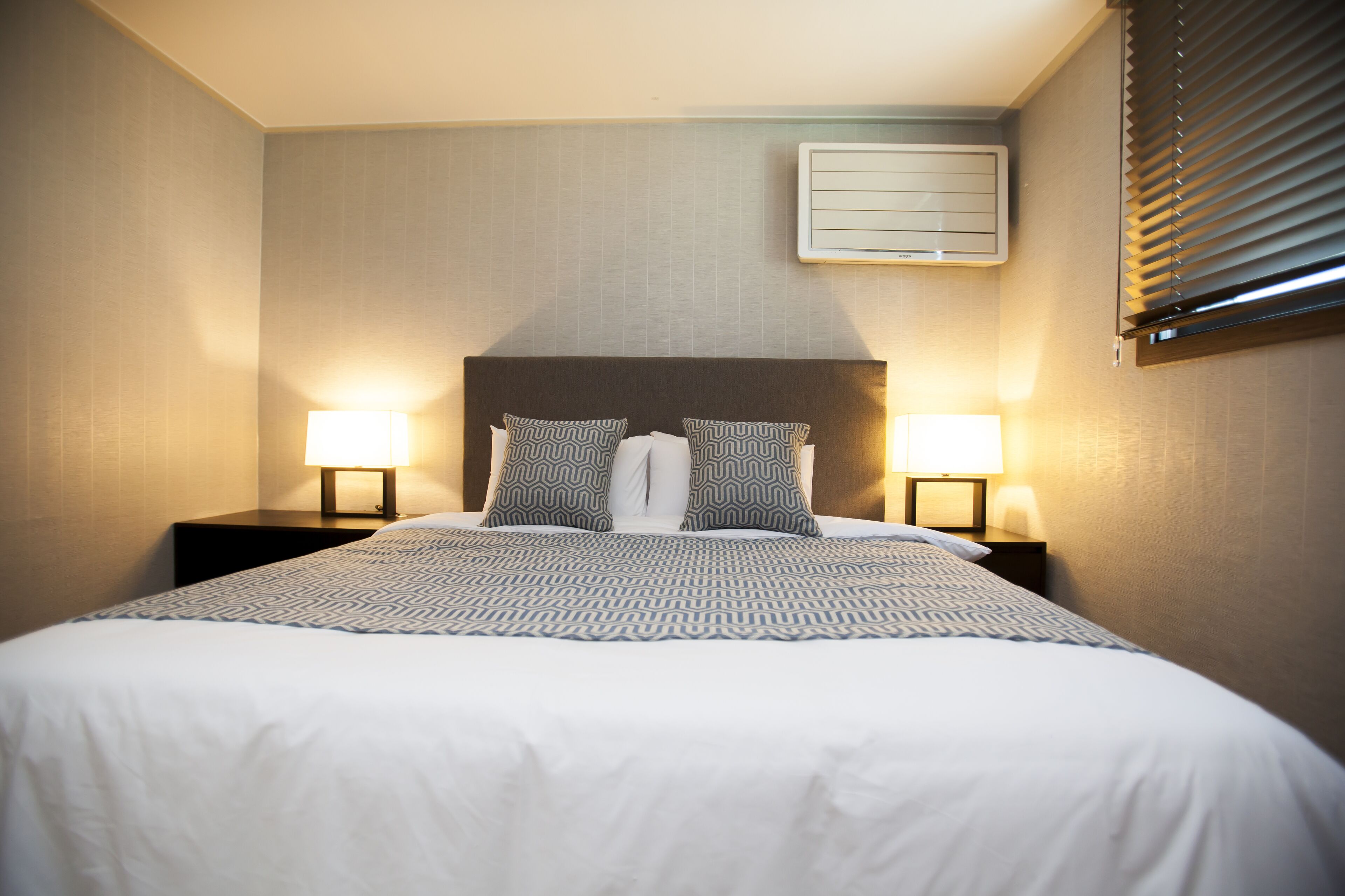 A neatly made bed with patterned pillows, two bedside lamps, and a wall-mounted air conditioner.