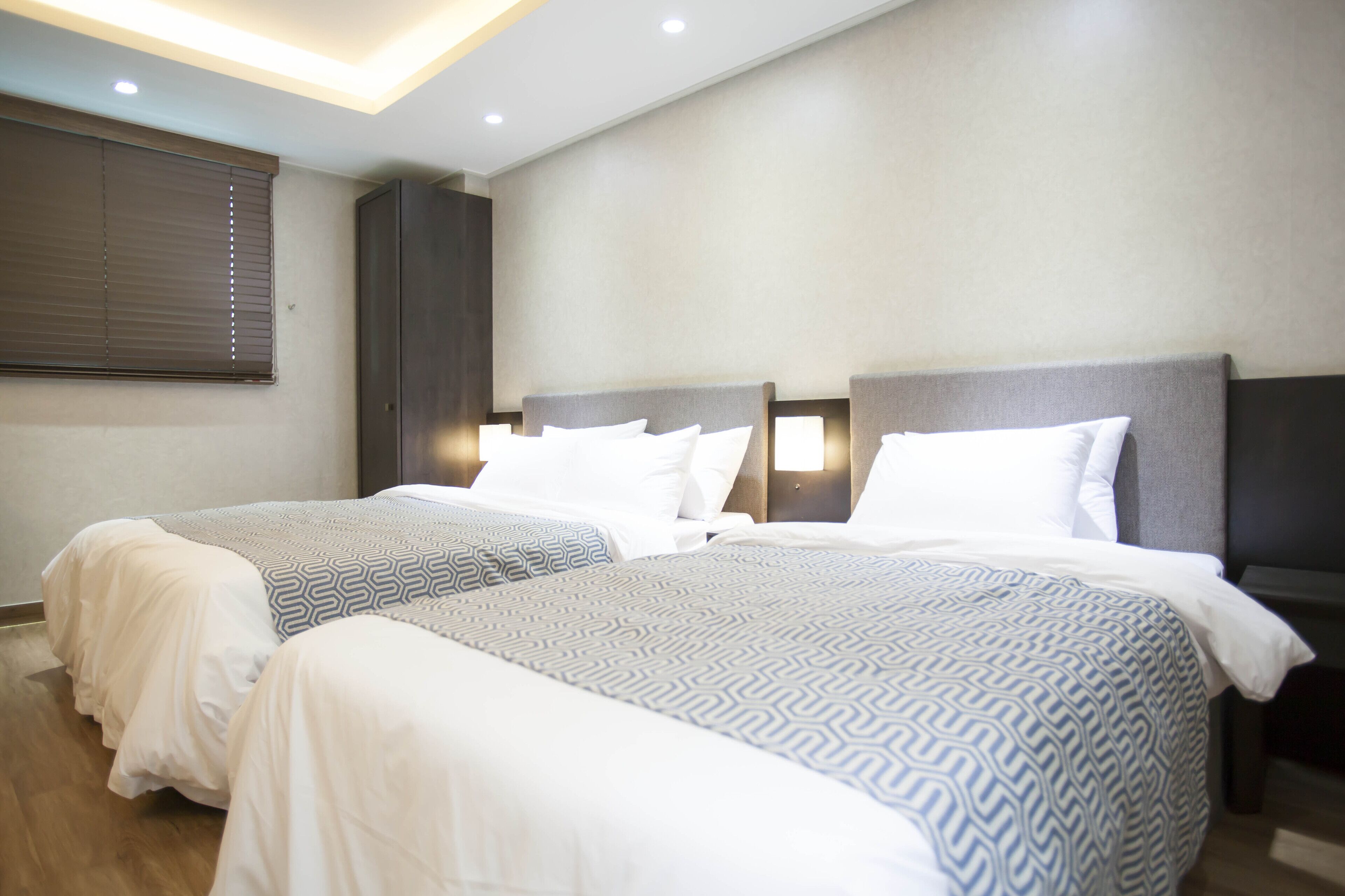 Habitación de hotel con dos camas, cada una con cabecera, ropa de cama blanca y una colcha estampada. Hay un armario y una mesita de noche con lámpara.