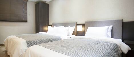 Habitación de hotel con dos camas, cada una con cabecera, ropa de cama blanca y una colcha estampada. Hay un armario y una mesita de noche con lámpara.
