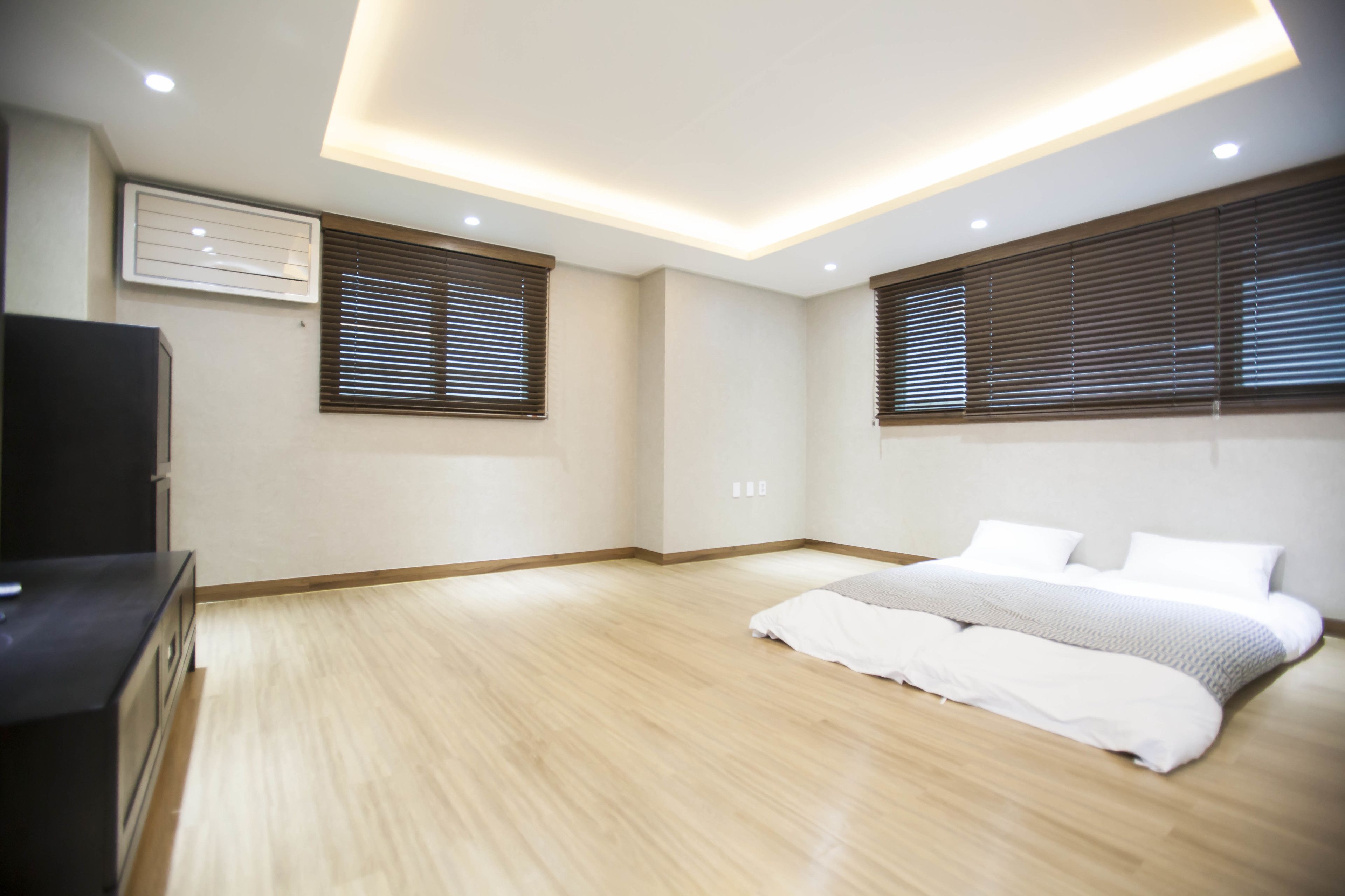A bedroom with a wooden floor, a bed with white bedding, a black TV stand, and a ceiling light fixture.