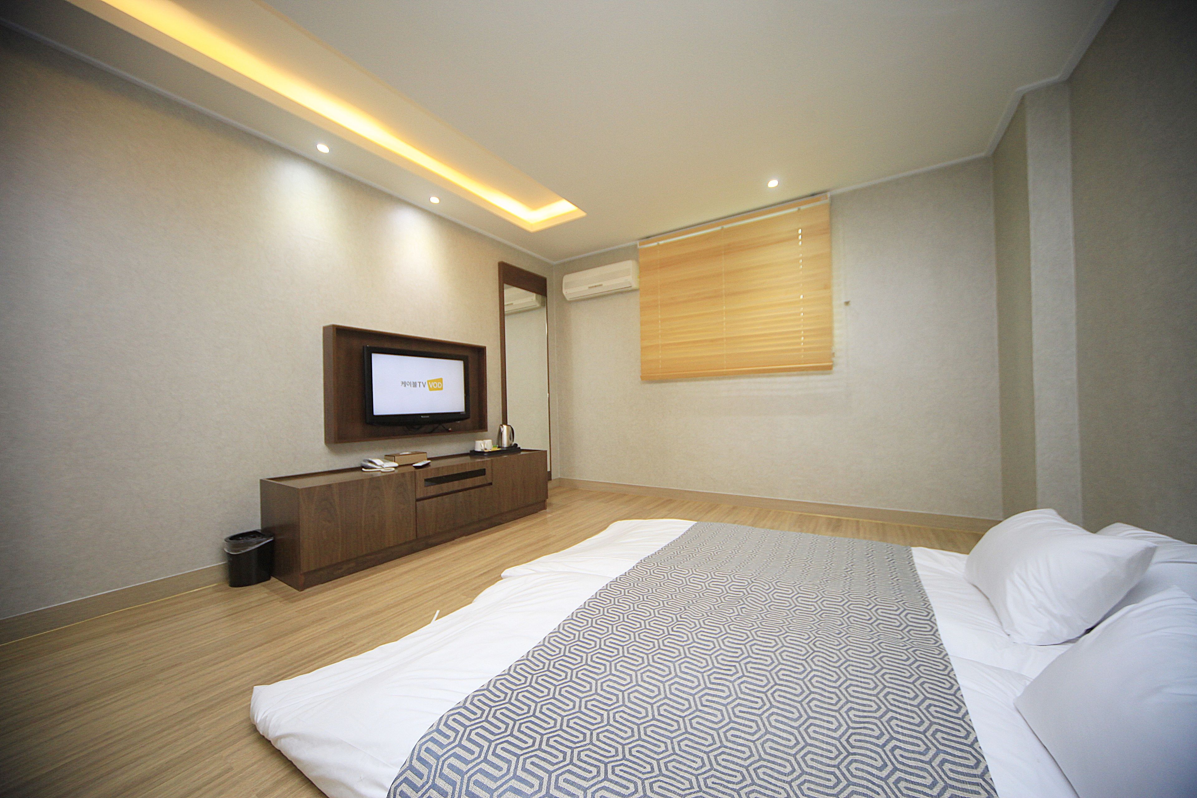 A modern hotel room with a large bed, a TV mounted on the wall, a wooden dresser, and a window with blinds.