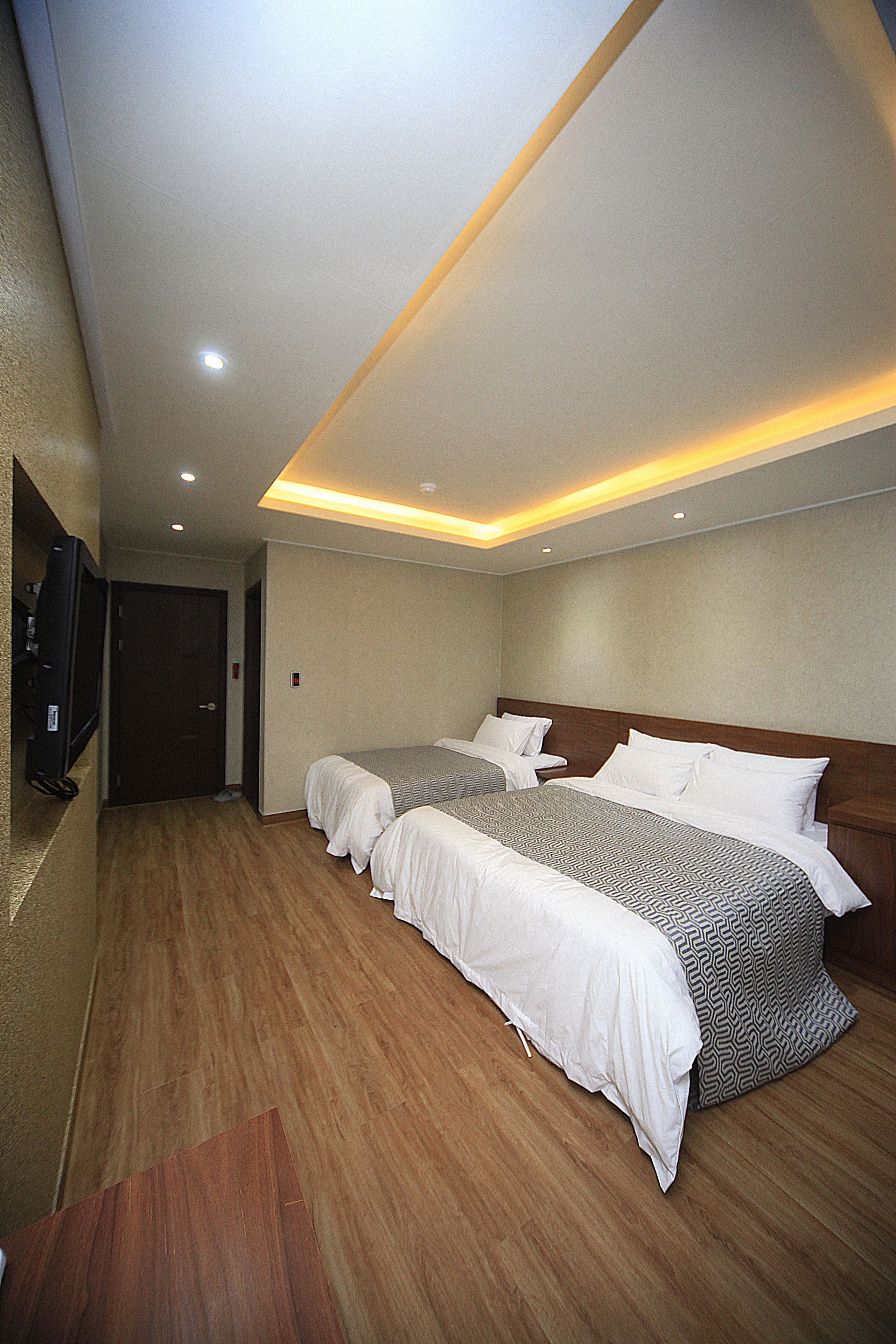 A hotel room with two beds, wooden flooring, a television, and recessed ceiling lighting.