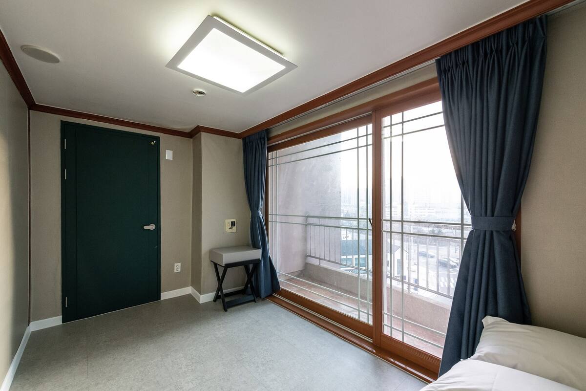 A hotel room with a bed, a small table, a bench, a window with curtains, and a door.