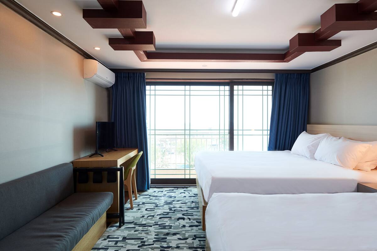 A hotel room with two beds, a sofa, a desk, and a large window with curtains.
