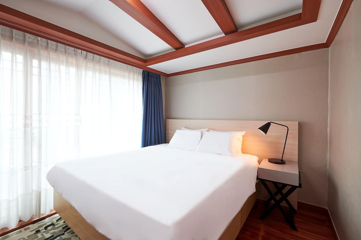 A hotel room with a large bed, a nightstand, a lamp, a window with sheer curtains, and a wooden ceiling.