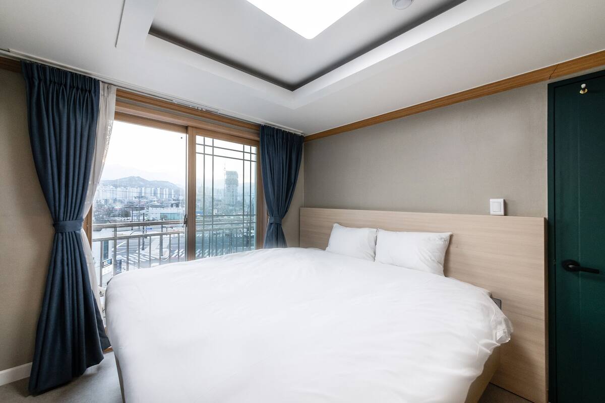 A hotel room with a large bed, a skylight, a city view, and a green door.
