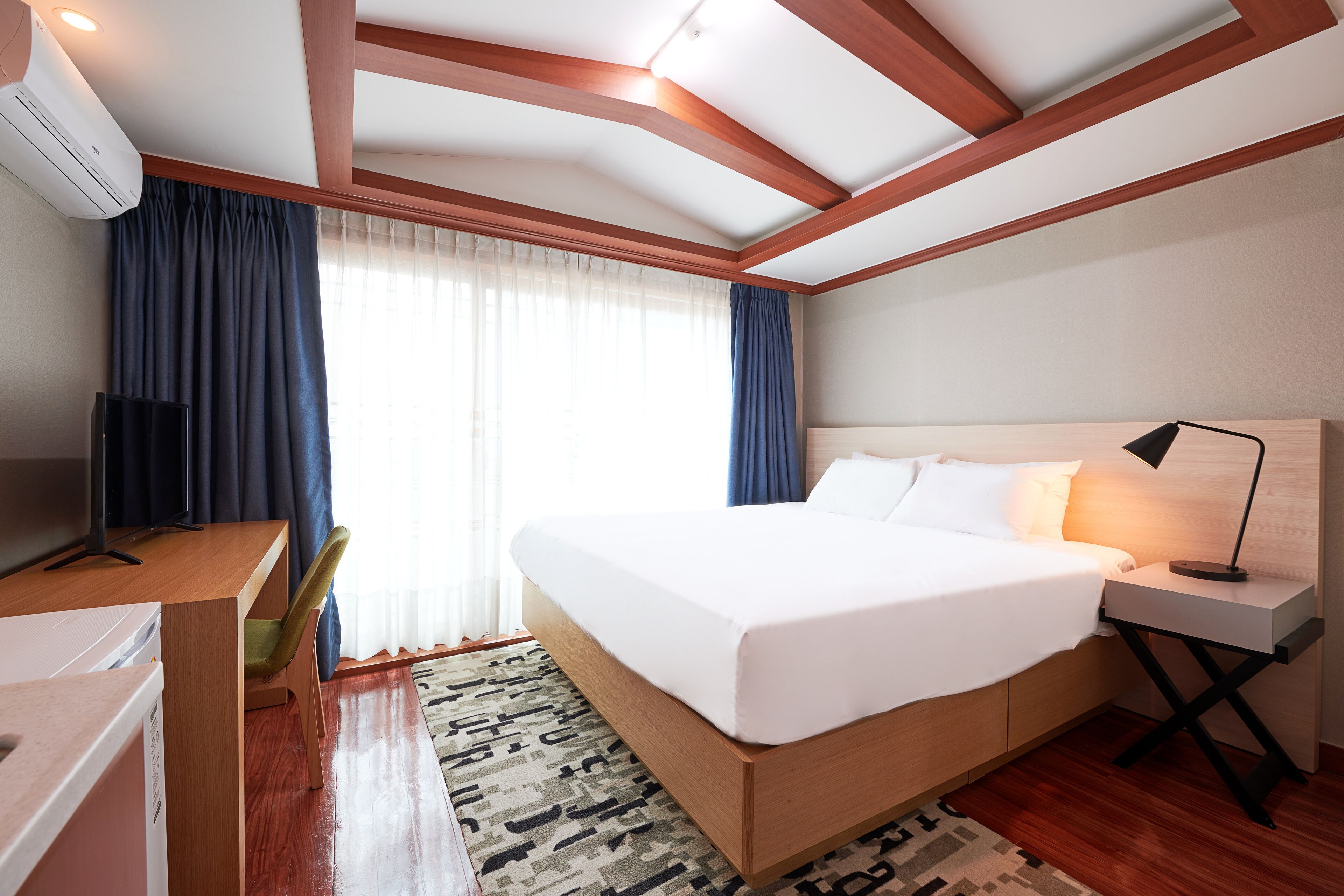 Habitación de hotel con cama, escritorio, silla, lámpara y aire acondicionado.