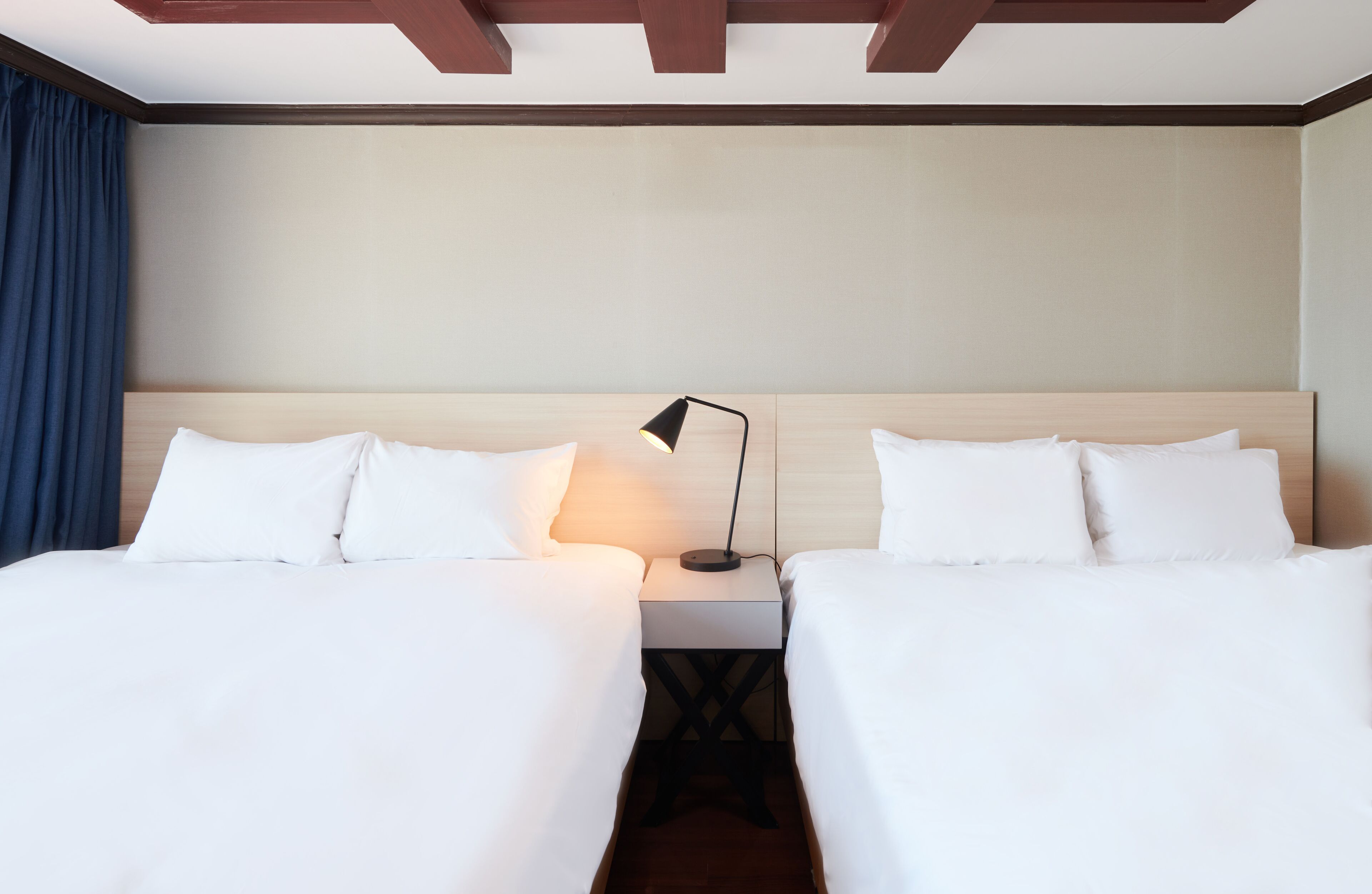A hotel room with two beds, a nightstand with a lamp, wooden ceiling beams, and a blue curtain.