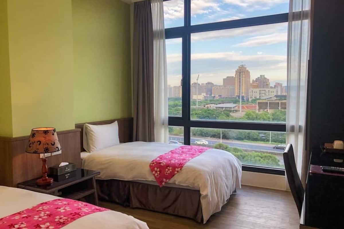 Hotelkamer met een groot raam, een bed met een roze bloemenprint, een nachtkastje met een lamp en uitzicht op een stadsbeeld.