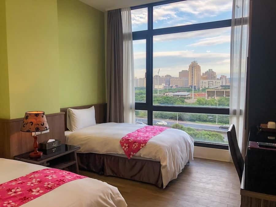 Ein Hotelzimmer mit großem Fenster, einem Bett mit blumigem Rosa-Bettüberzug, einem Nachttisch mit Lampe und Blick auf eine Stadtlandschaft.