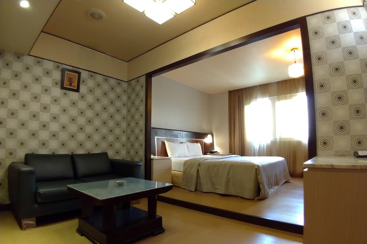 Een hotelkamer met een bank, een glazen tafel, een bed met hoofdeinde en een grote spiegel die de kamer weerspiegelt.