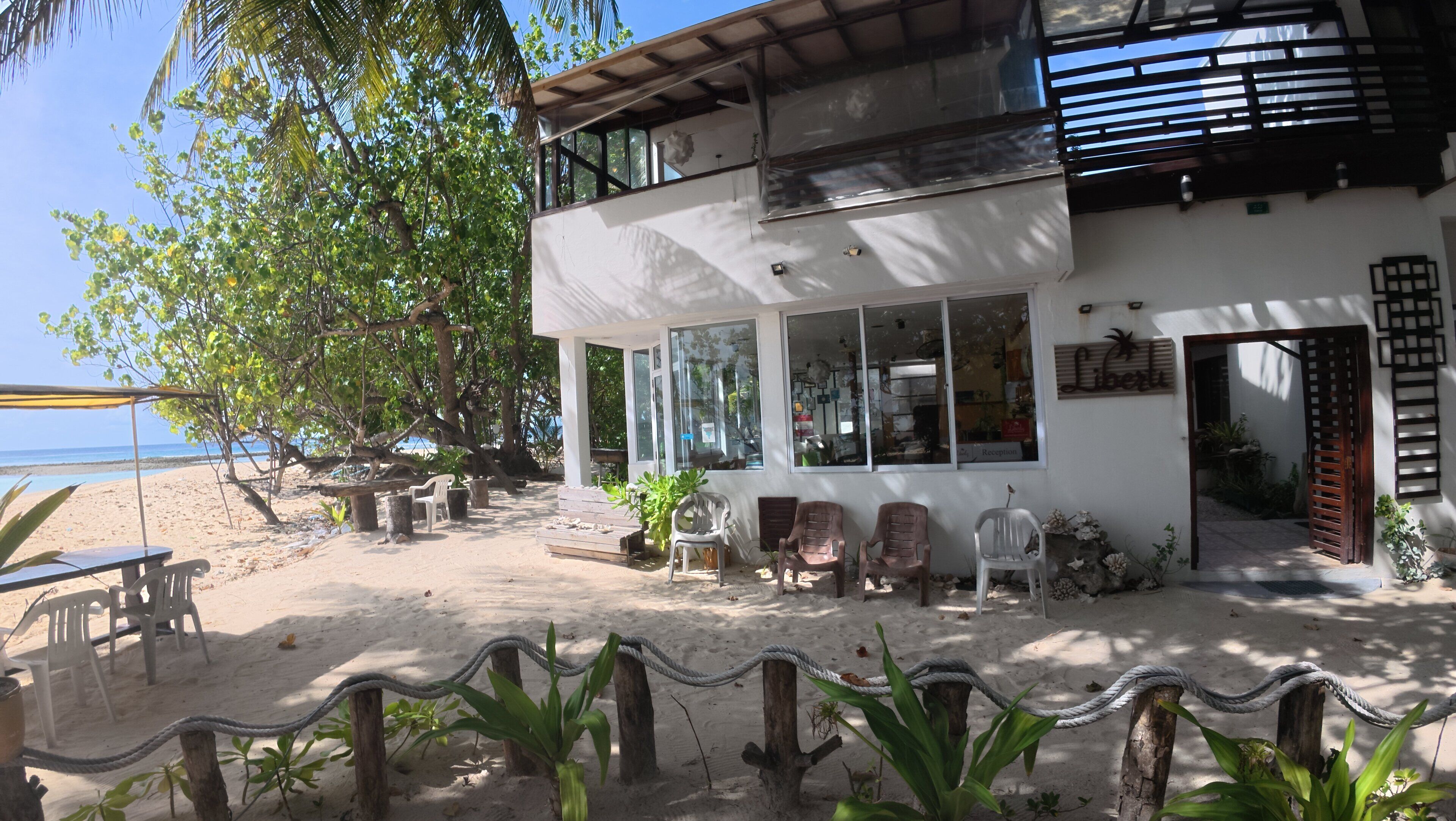 Una propiedad frente a la playa con un edificio de dos pisos, asientos exteriores y una zona arenosa con plantas.