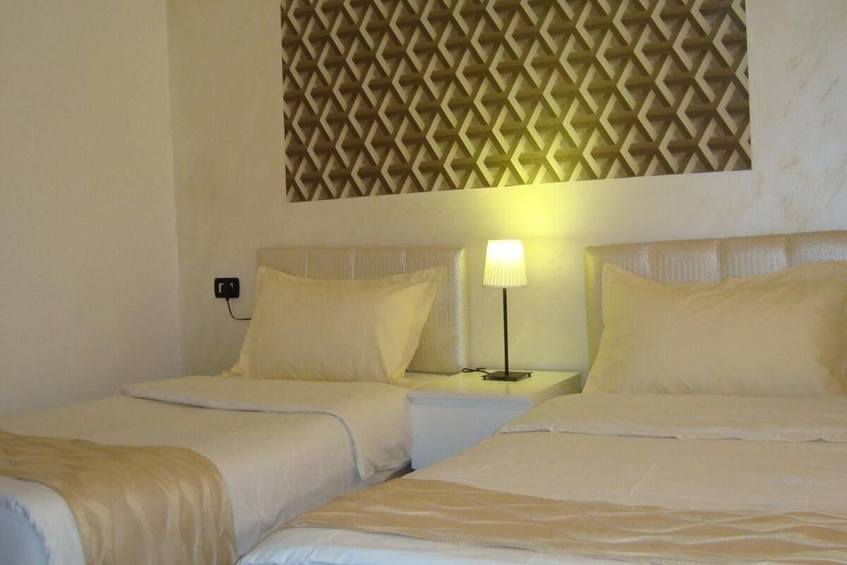 A hotel room with two beds, each with a headboard, a bedside lamp, and a power outlet on the wall.