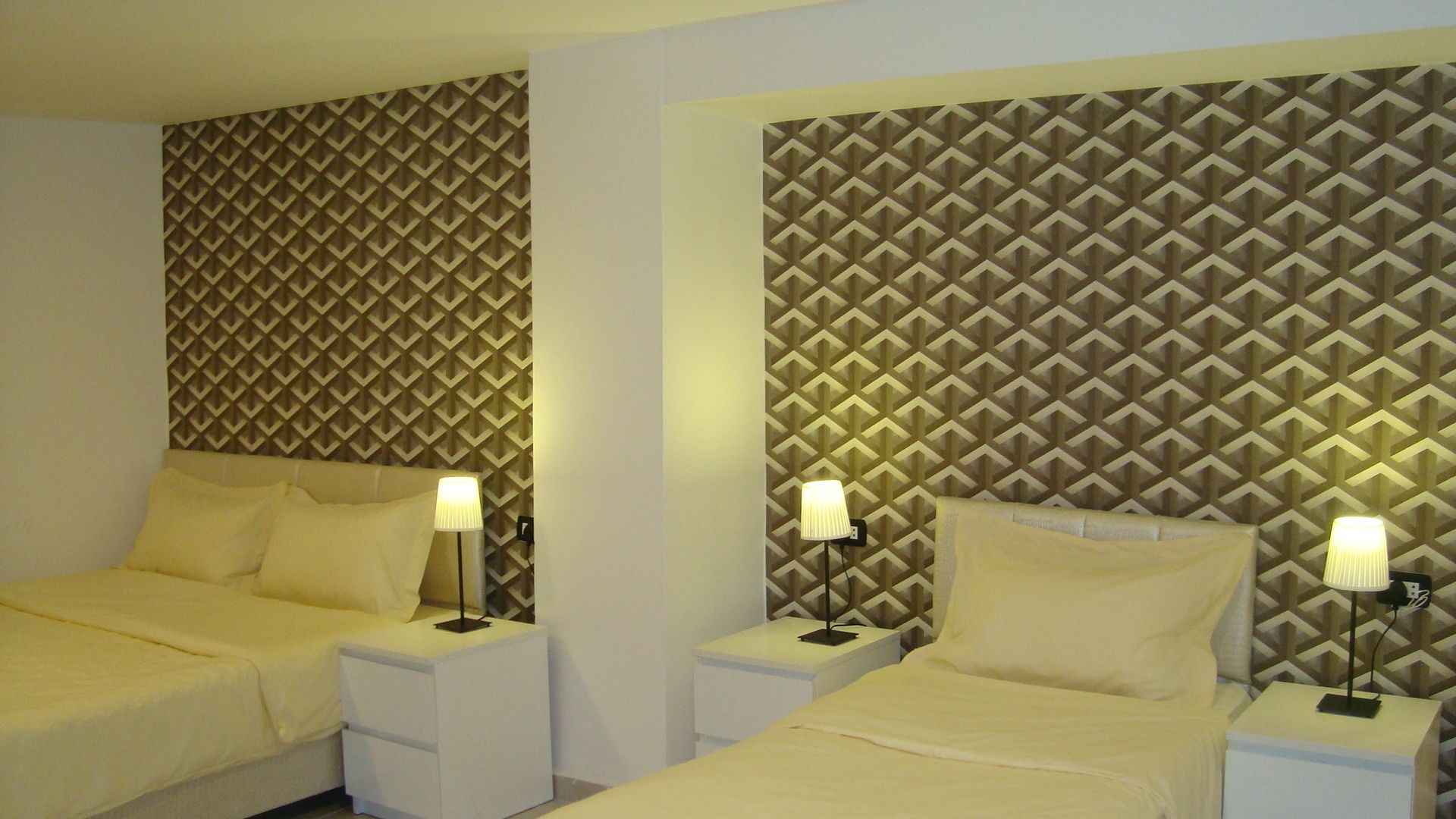 A hotel room with two beds, each with a nightstand and a lamp. The walls have a geometric patterned wallpaper.