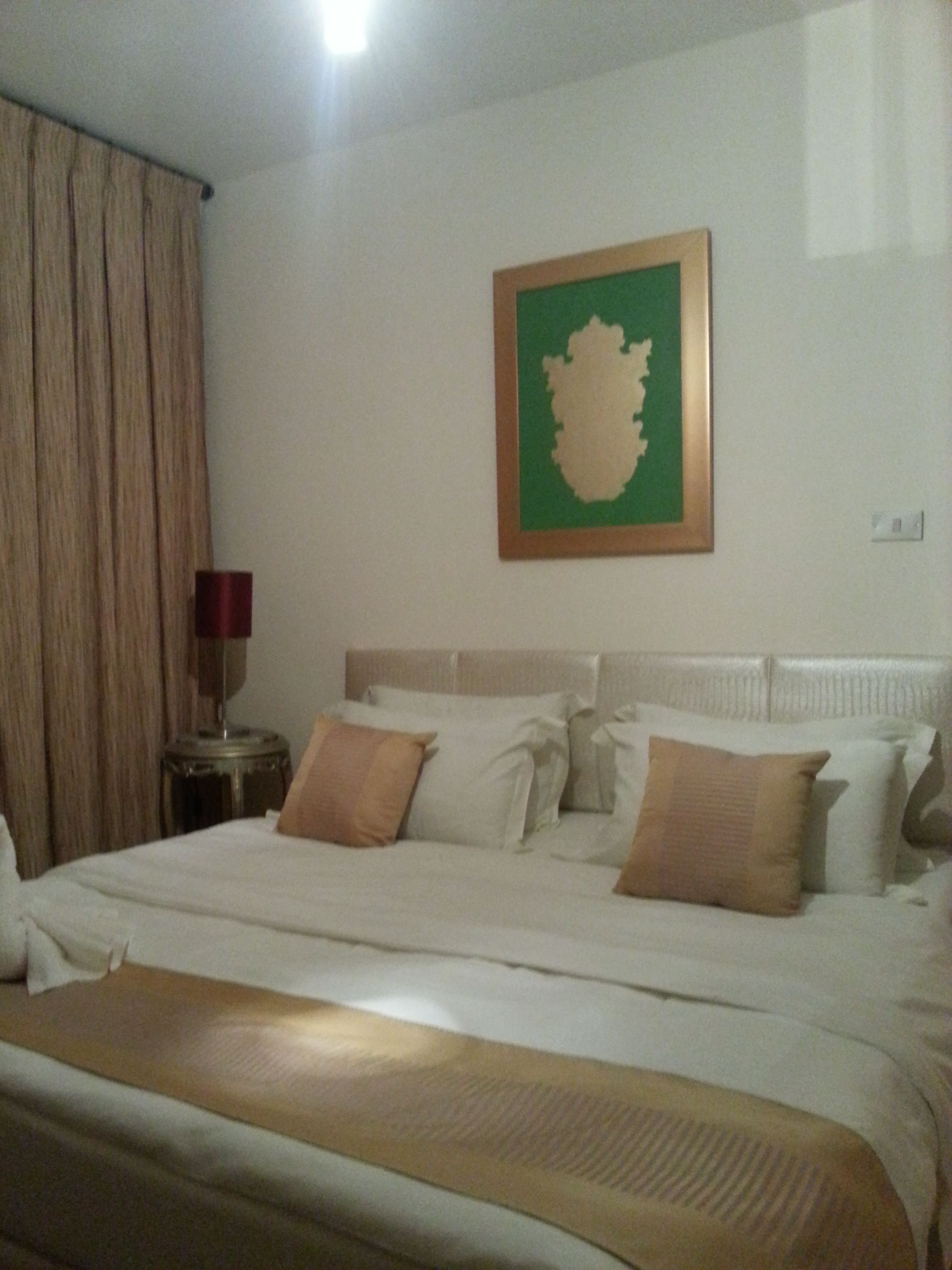 A hotel room with a large bed, a nightstand with a lamp, and a green-framed artwork on the wall.