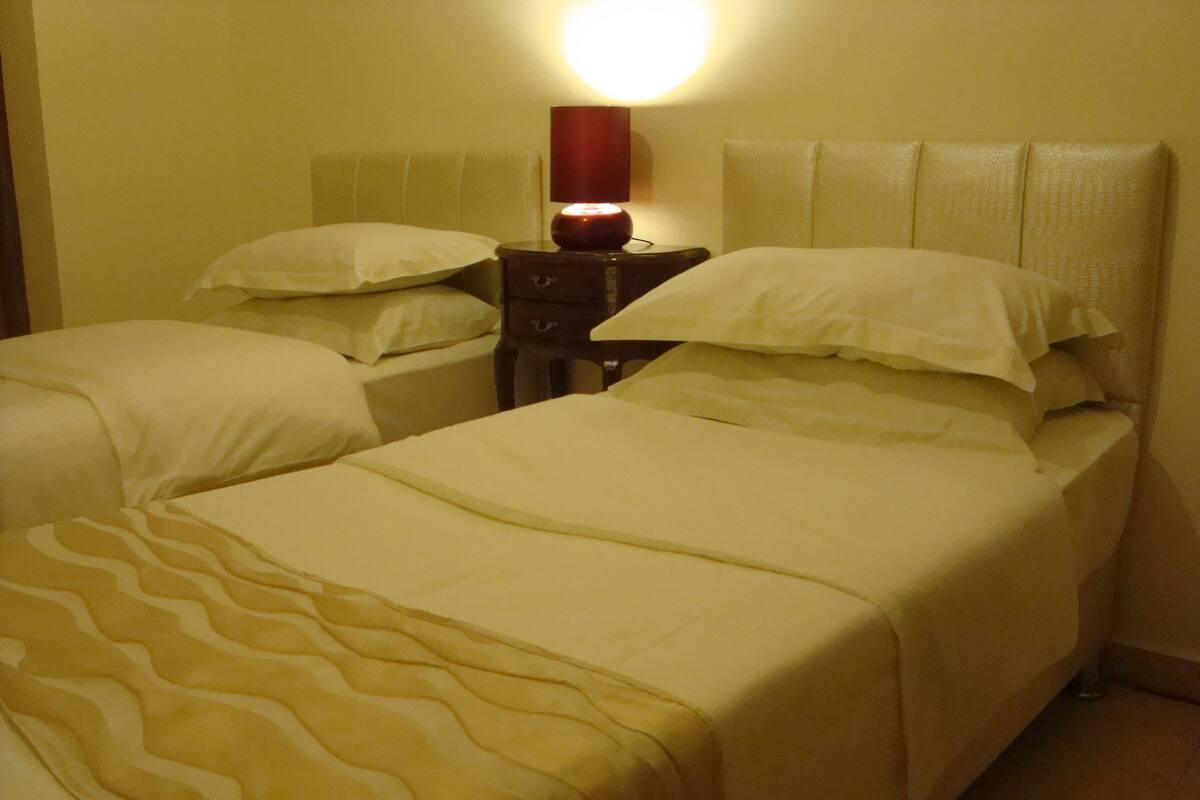 A hotel room with two beds, a nightstand with a lamp, and a wall-mounted light.