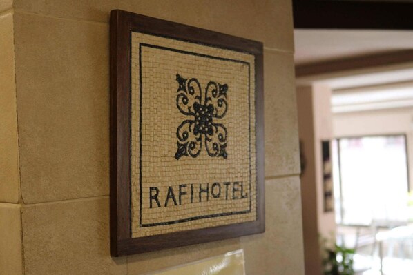 A framed sign with a decorative design and the text "RAFI HOTEL" on a beige wall.