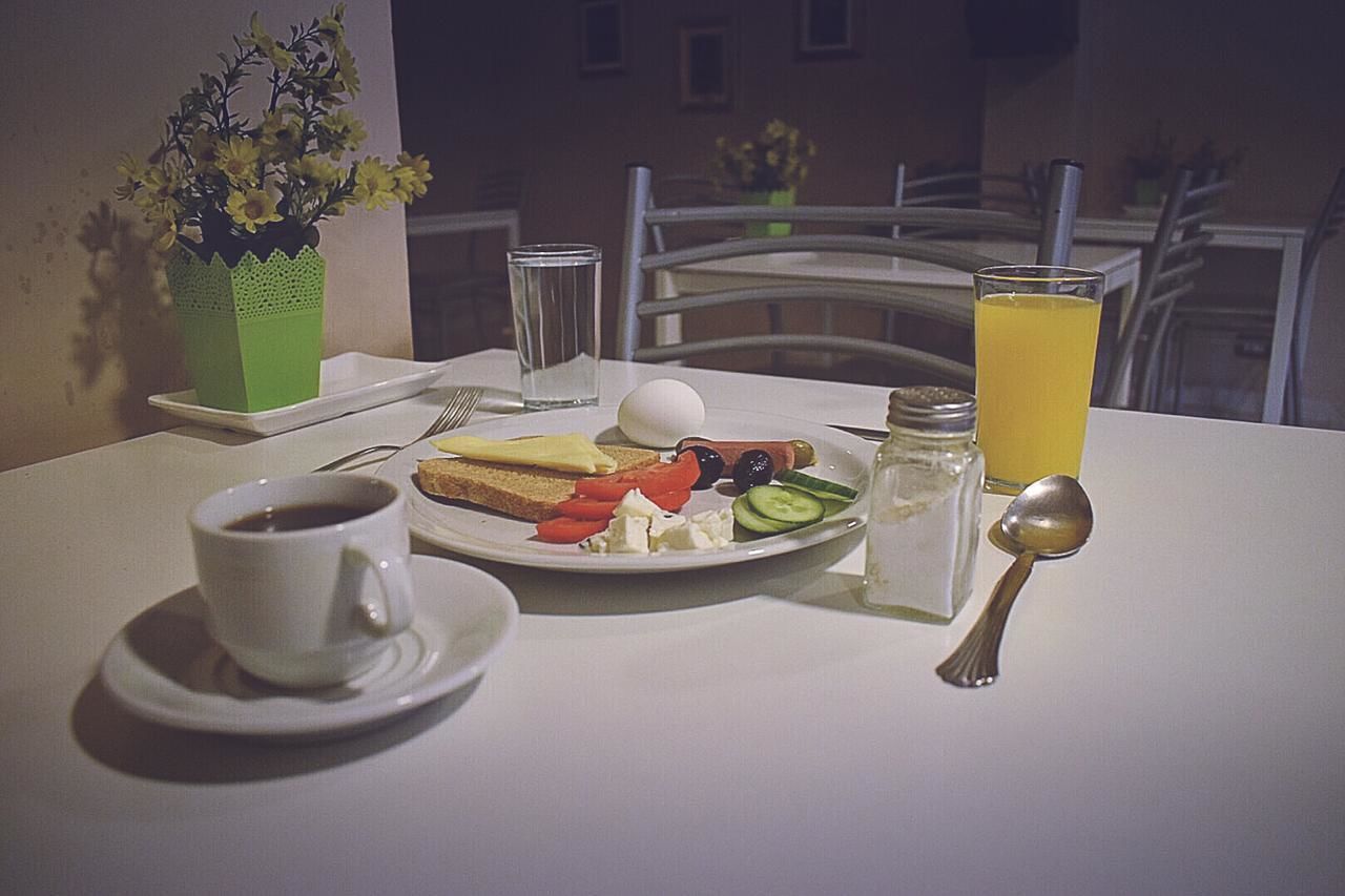 A breakfast table with a cup of coffee, a plate of food including eggs, tomatoes, cucumbers, olives, and cheese, a glass of orange juice, and a small vase with flowers.