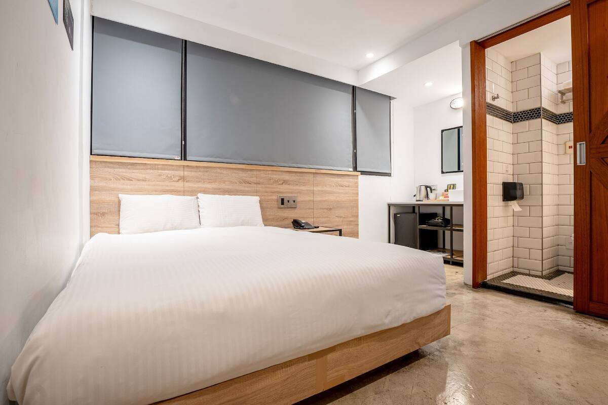 A modern hotel room with a large bed, a wooden headboard, a bedside table with a phone, and a shower area with a tiled wall and wooden door.