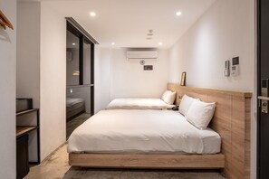 A modern hotel room with two beds, a built-in wardrobe, an air conditioning unit, and a small desk area.