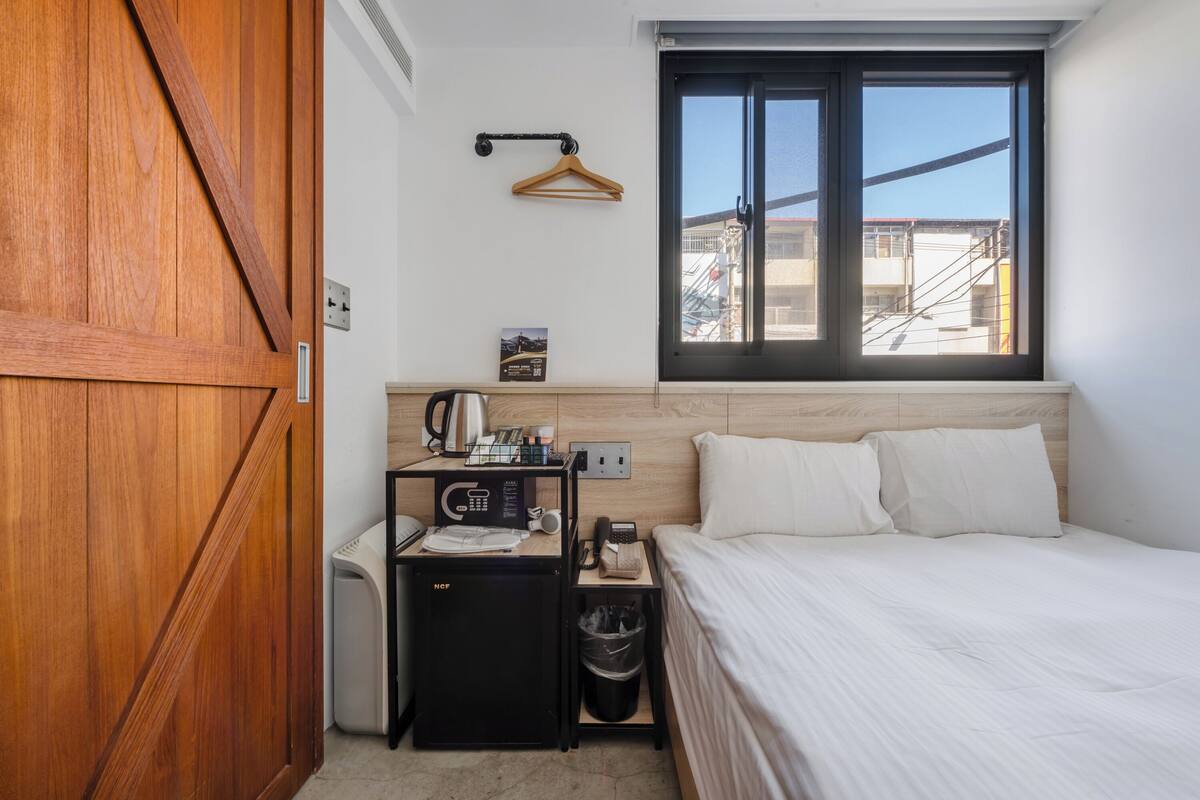 A hotel room with a wooden wardrobe, a compact kitchenette, a bed with white bedding, a window with a view of buildings, and a hanging coat rack.