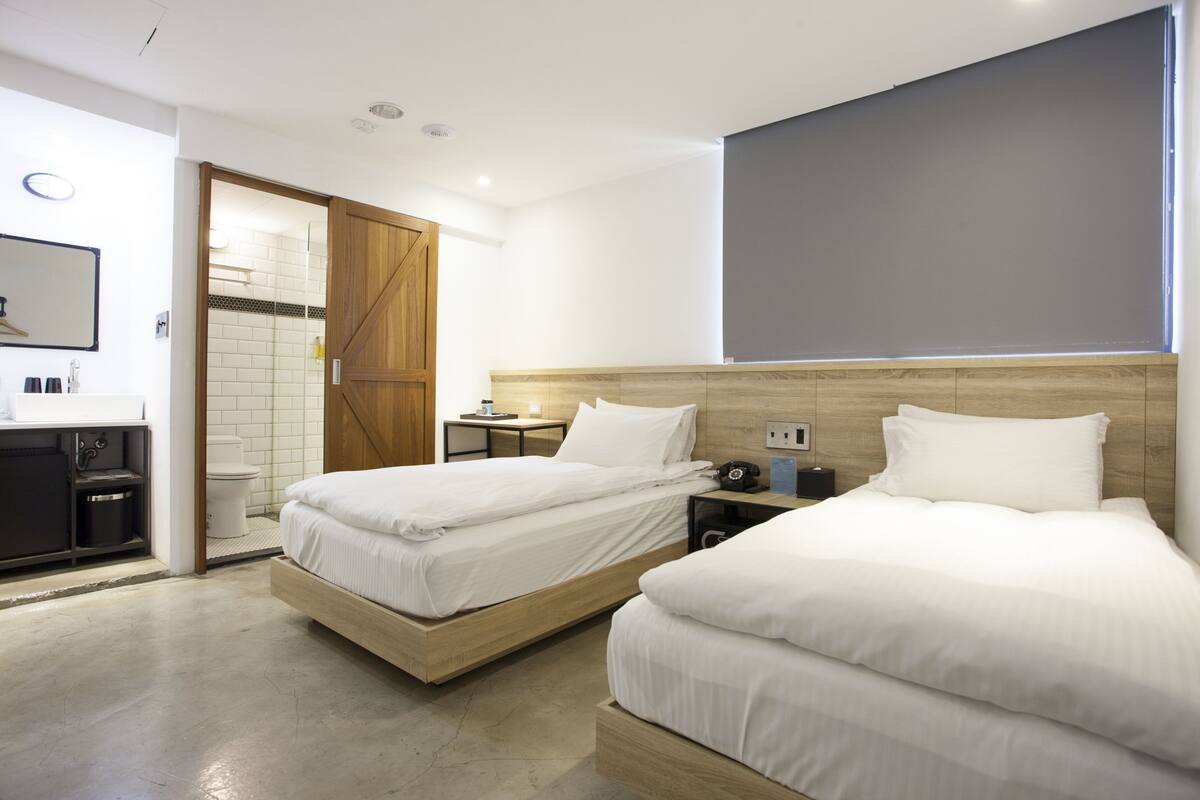 A hotel room with two beds, a wooden headboard, a small bedside table, a nightstand, a mirror, a wardrobe, and a bathroom with a toilet and sink.