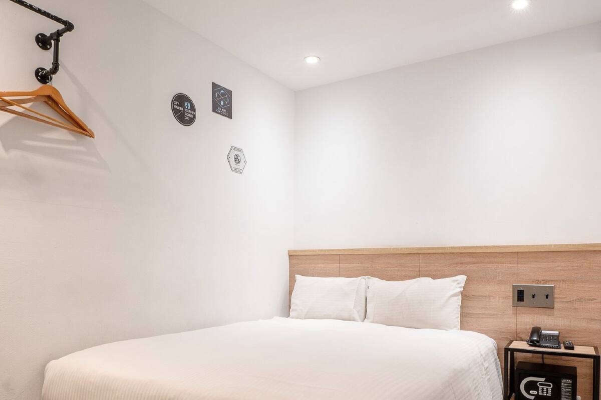 A modern bedroom with a wooden headboard, a bed with white linens, a bedside table with a phone, and a wall-mounted coat rack.