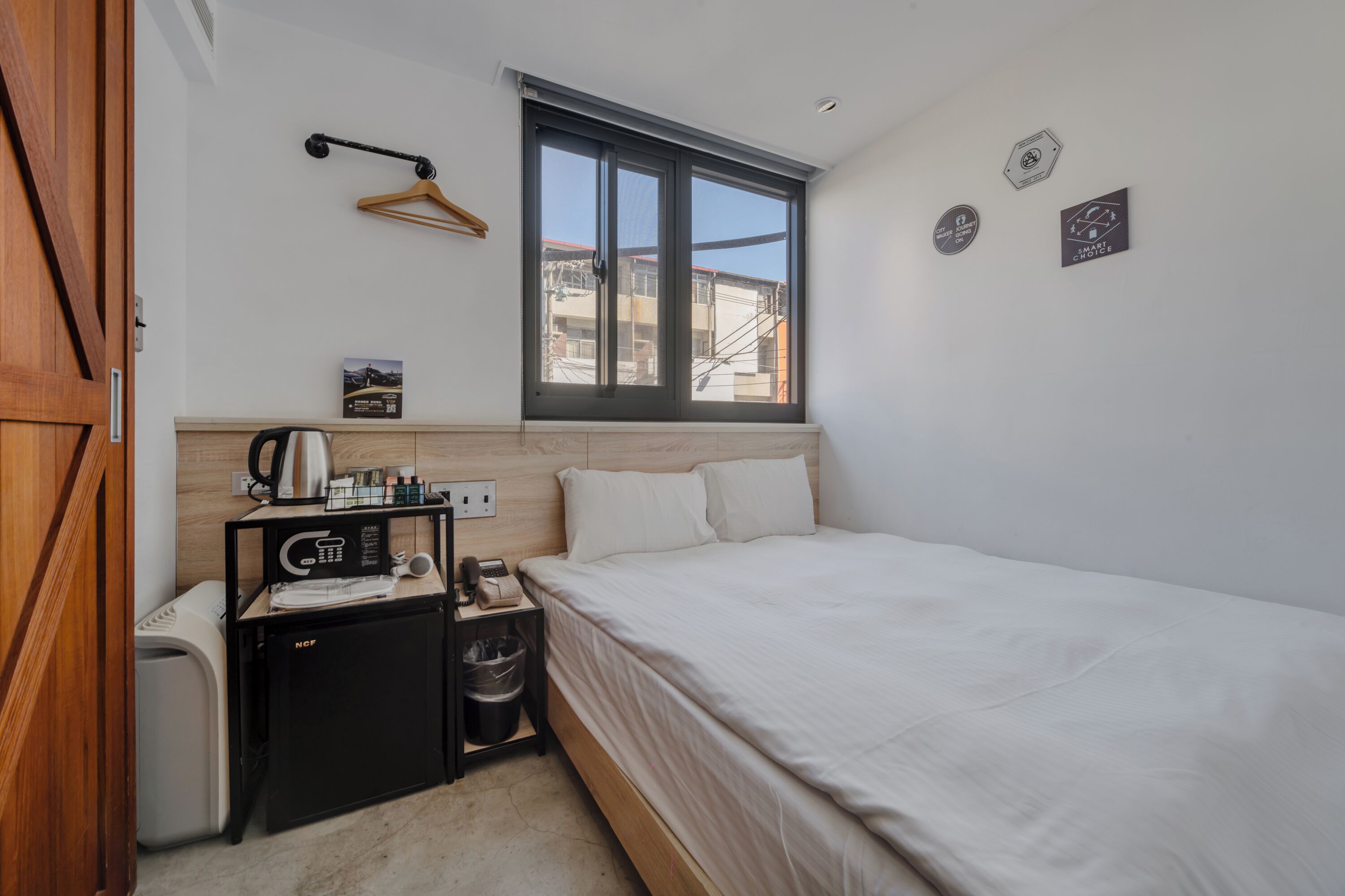 A bedroom with a bed, a small desk with a coffee machine, a hanging light, a window, and a wall-mounted heater.