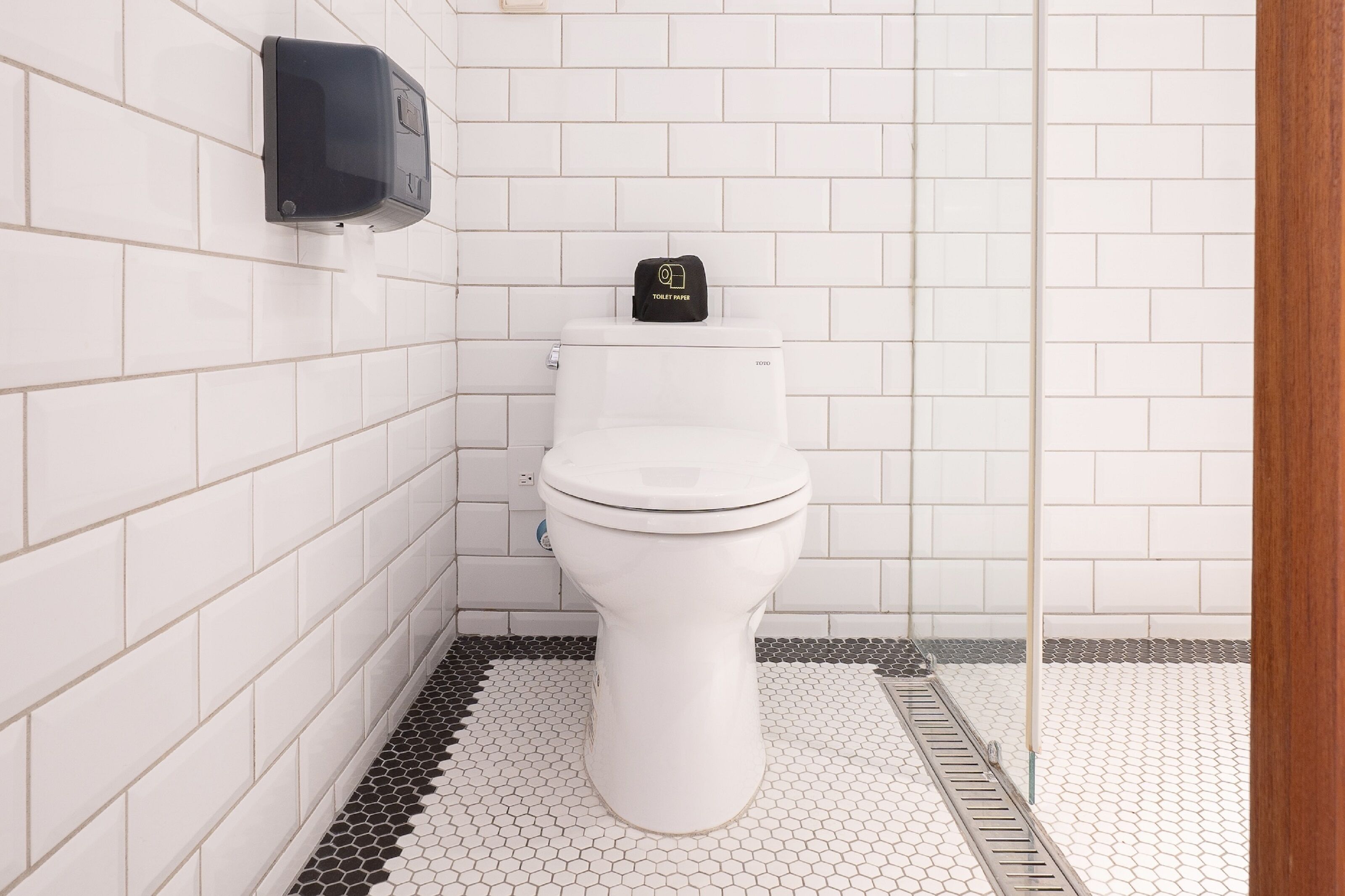 Eine weiße Toilette mit schwarzem Deckel, ein wandmontierter Seifenspender und ein Boden mit sechseckigem Muster.