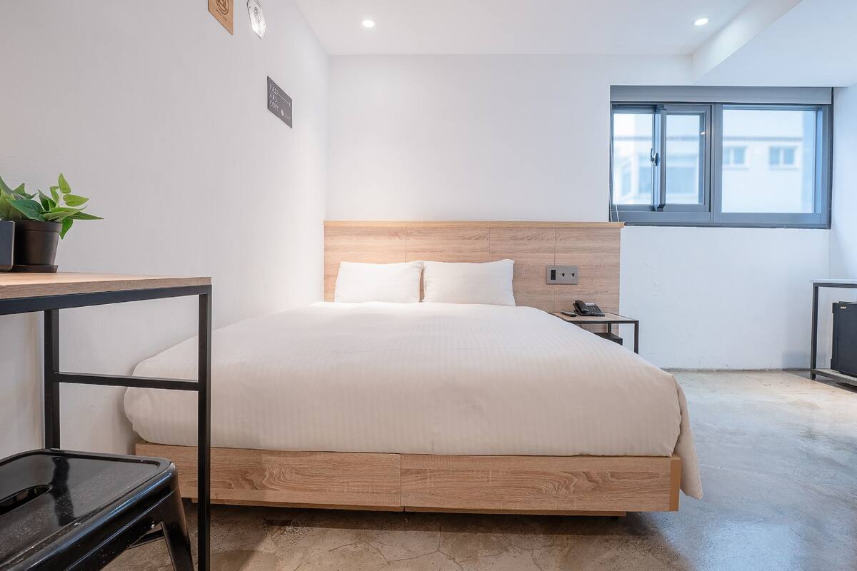 A modern bedroom with a wooden headboard, a single bed, a bedside table with a phone, a small desk, and a potted plant.