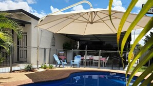 A swimming pool with a patio area, including a covered seating area with chairs and a table, and a fenced garden.