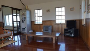Una habitación con piso de madera, una mesa, una silla, un refrigerador, un televisor y dos ventanas.