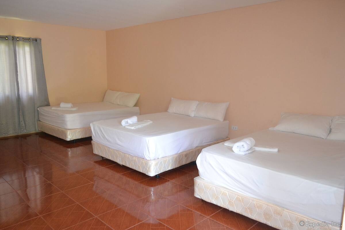 A hotel room with three single beds, each with a white mattress and pillow, and a bathroom with a shower curtain.