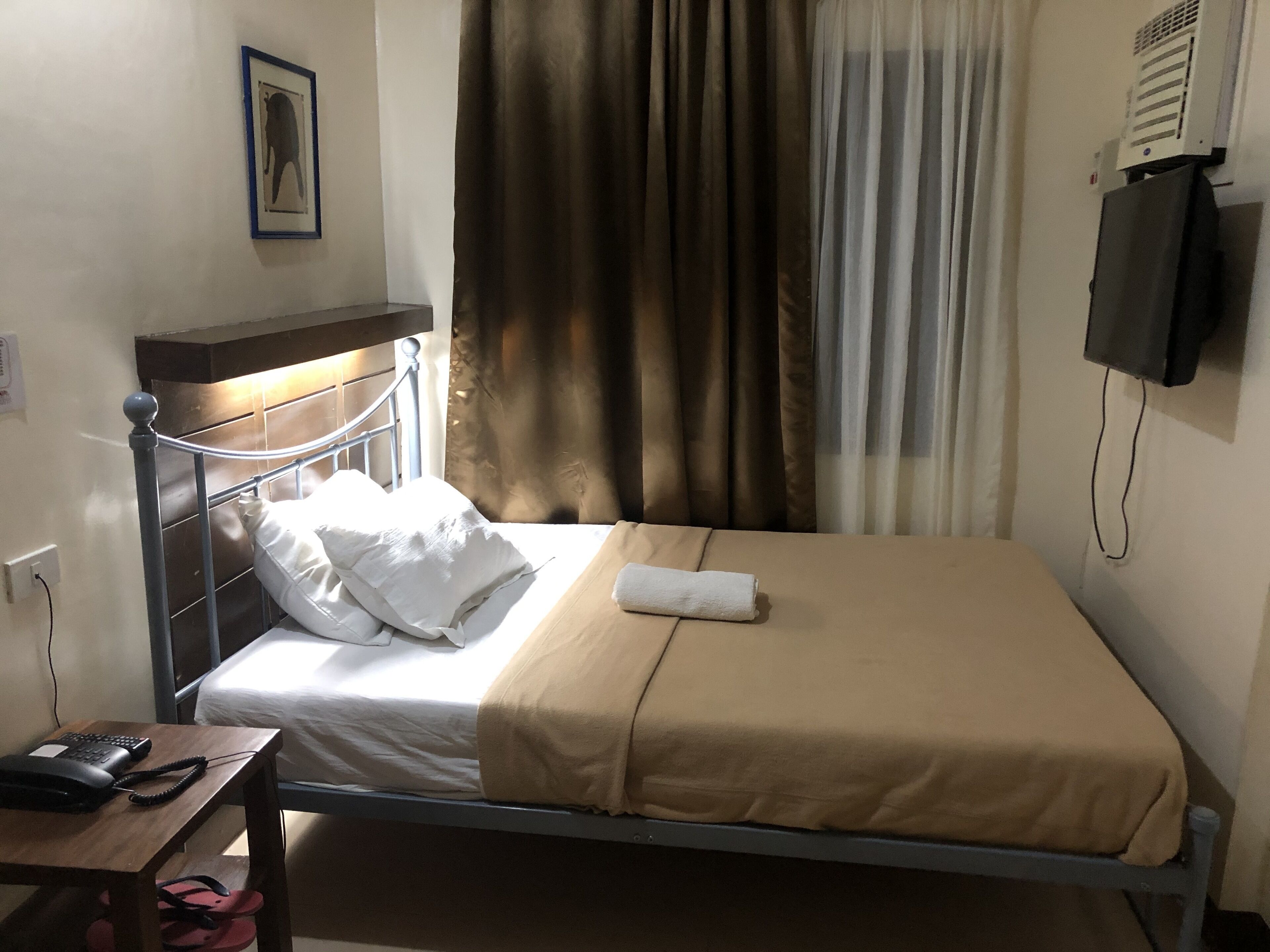 Una cama individual con cabecera, una mesita de noche con teléfono y chanclas, un televisor montado en la pared y una ventana con cortinas.