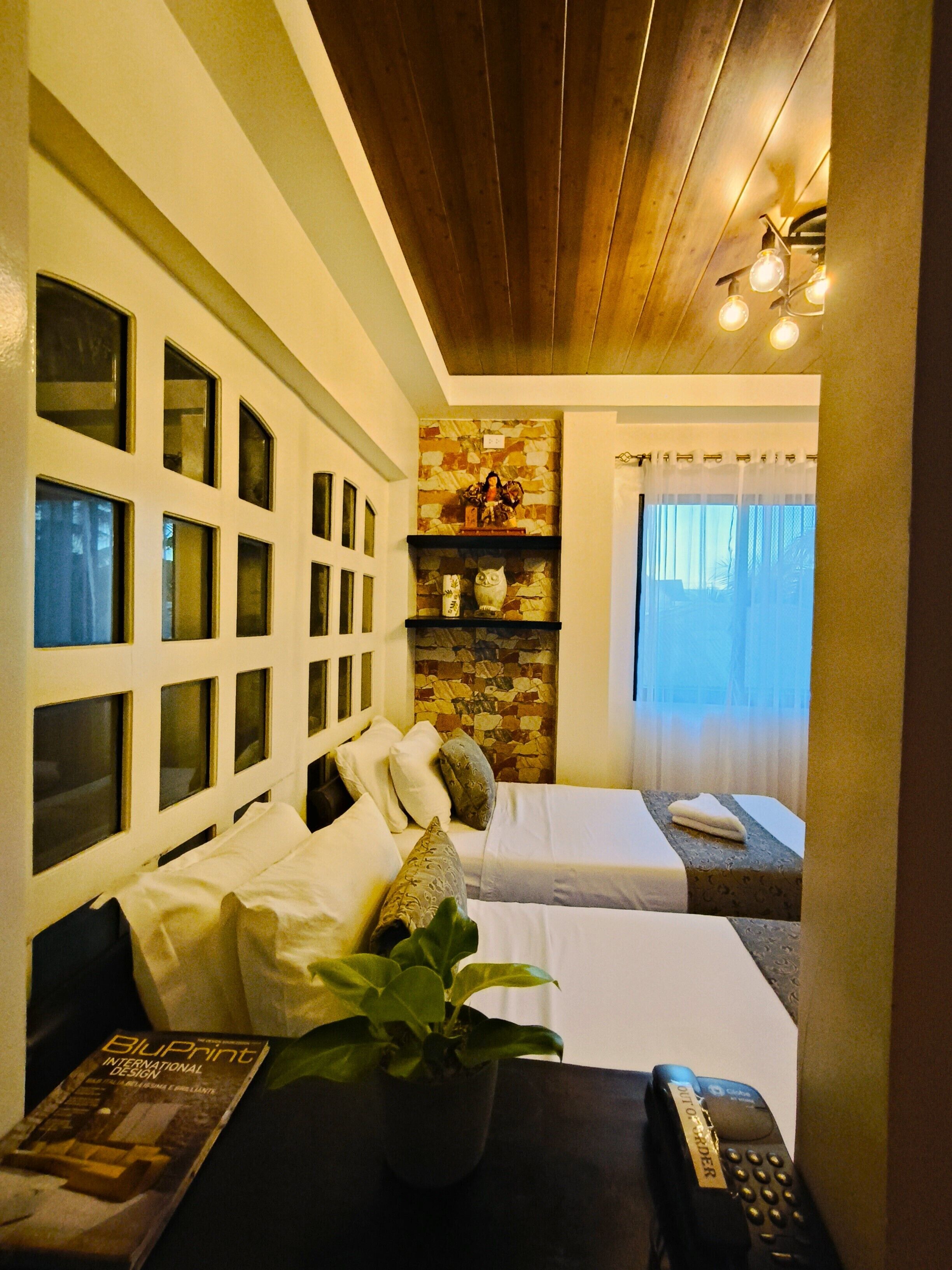 A hotel room with two beds, a stone wall with shelves, a window with curtains, and a remote control on the table.