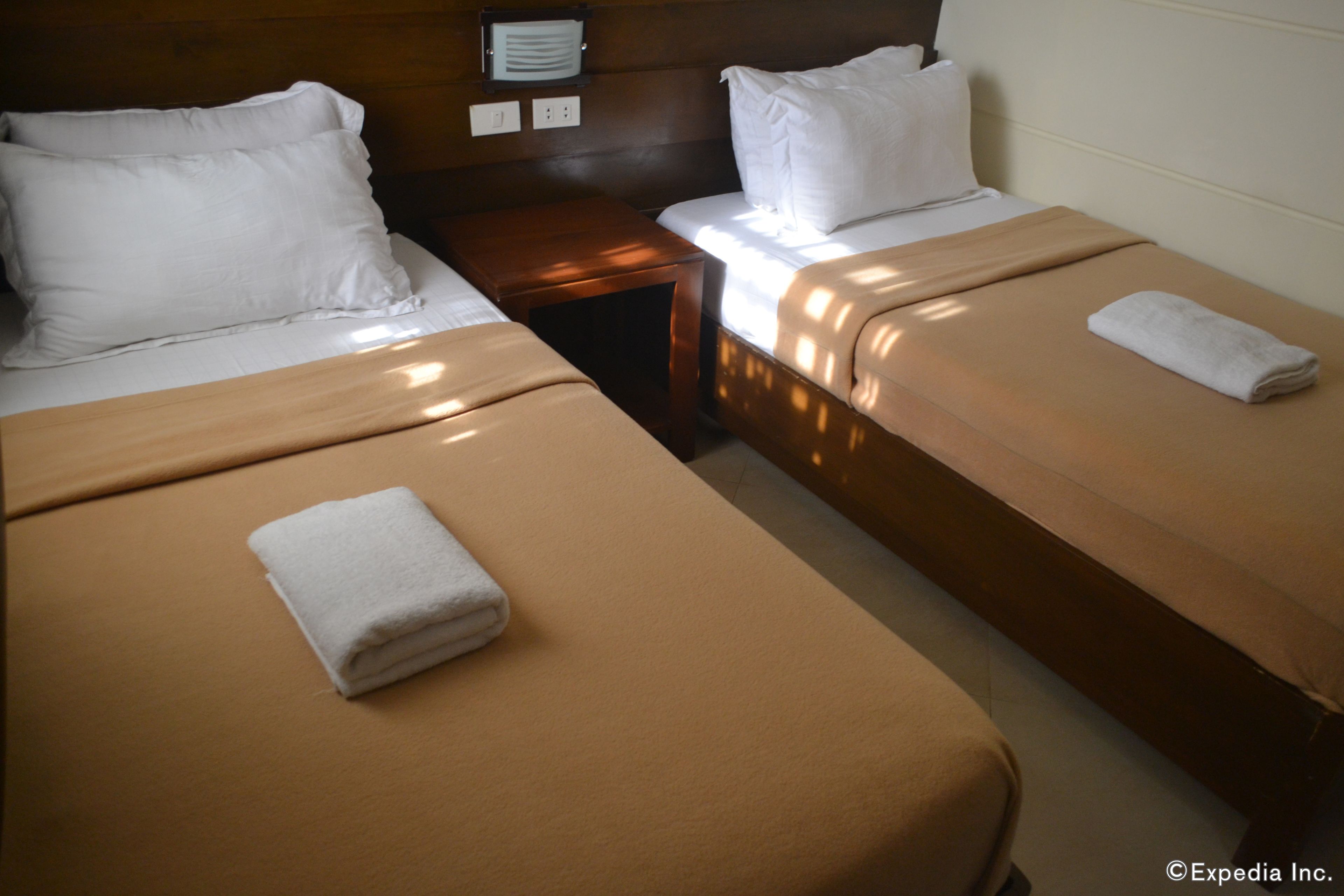 A hotel room with two single beds, each with a white pillow and a beige blanket. There is a wooden nightstand between the beds with a small lamp.
