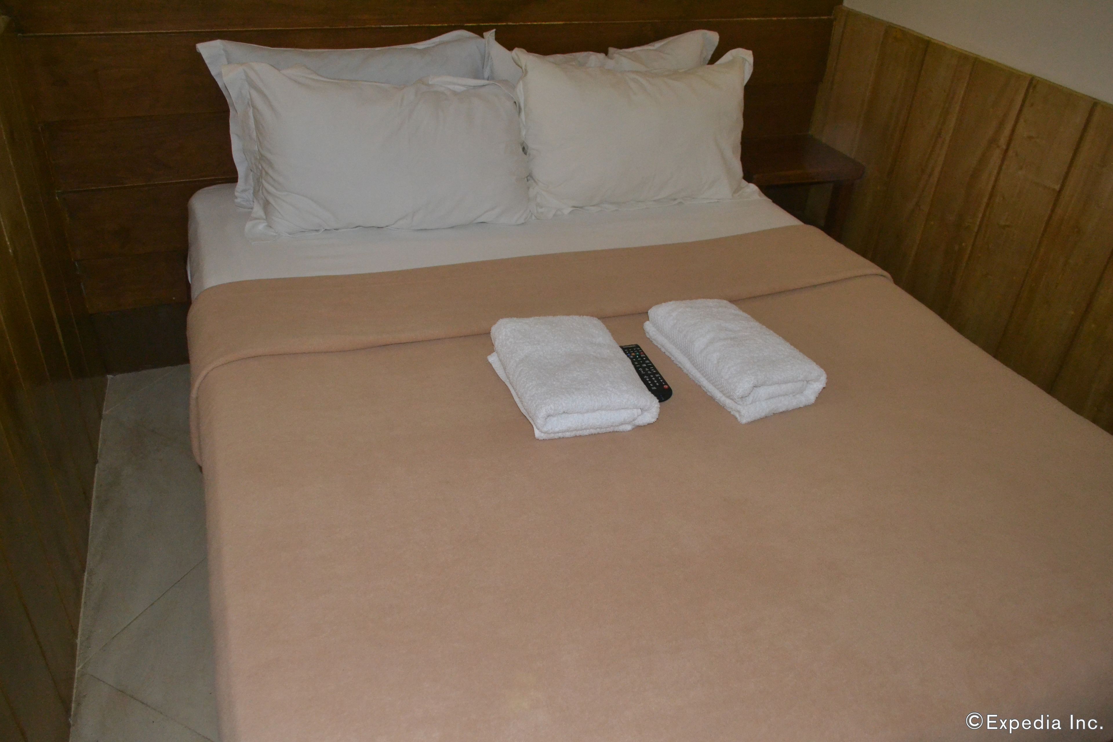 Una cama bien hecha con almohadas blancas y una manta color beige, un control remoto y dos toallas dobladas sobre la cama.