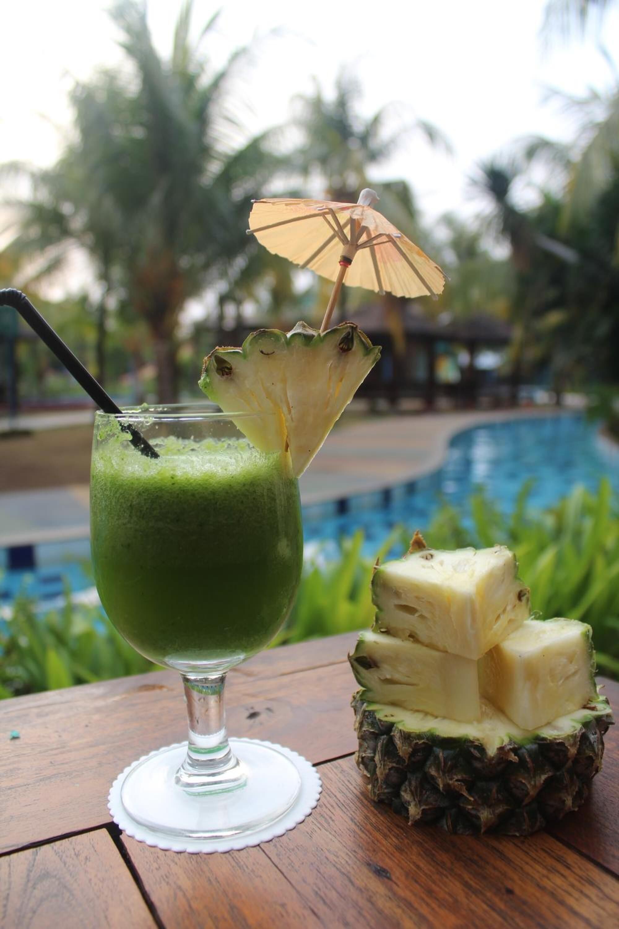 Un vaso de bebida verde con una decoración de piña y un paraguas sobre una mesa de madera cerca de una piscina.