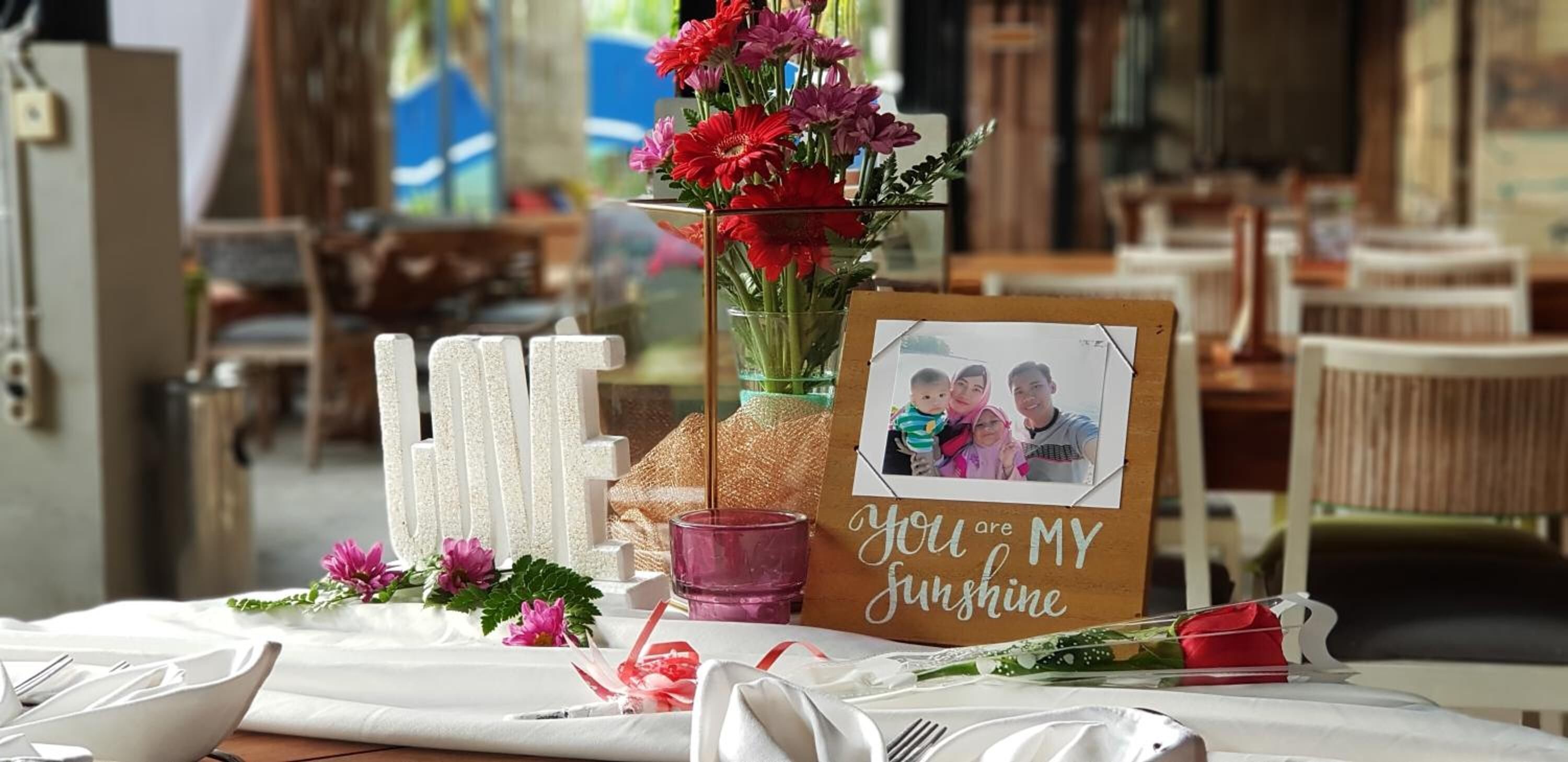 Une table dressée avec des nappes blanches, un vase en verre contenant des fleurs rouges, un cadre photo avec un message et une rose rouge.
