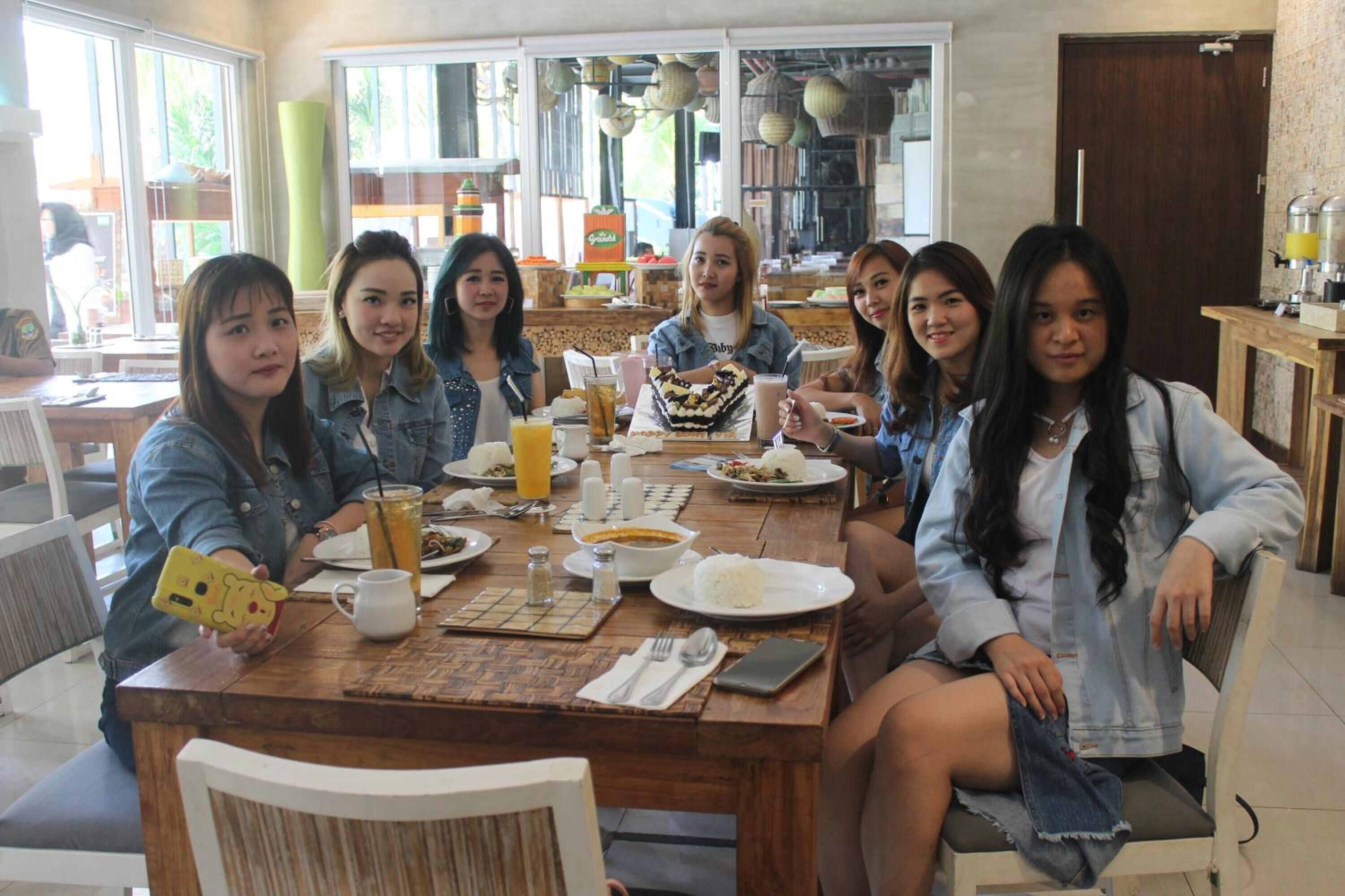 A group of people sitting around a wooden dining table with various dishes, drinks, and a smartphone on it.