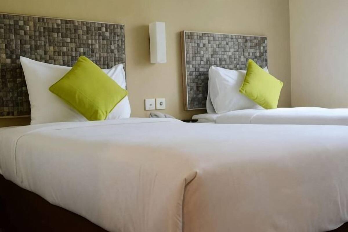 A hotel room with two beds, each with white bedding and a green pillow. There are wall-mounted light fixtures and a telephone on the nightstand.