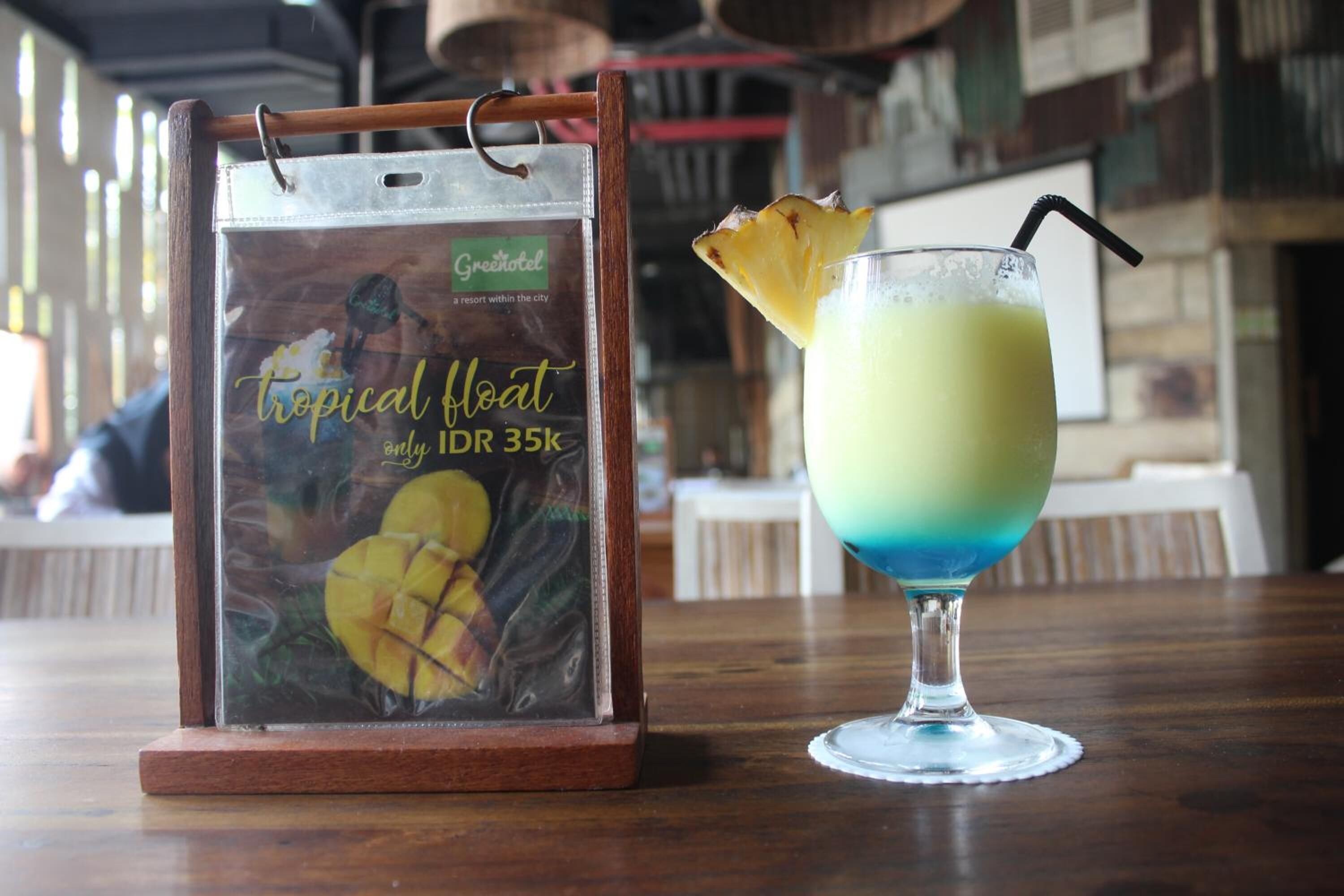 Bebida con temática tropical, adornada con piña, y un cartel de 'Greenhotel' que muestra una bebida flotante tropical y un precio de IDR 35k.