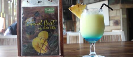 Bebida con temática tropical, adornada con piña, y un cartel de 'Greenhotel' que muestra una bebida flotante tropical y un precio de IDR 35k.