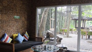 A room with a glass-top coffee table, a sofa with colorful cushions, and a view of an outdoor patio with tables and chairs. - Greenotel Cilegon (Cilegon)